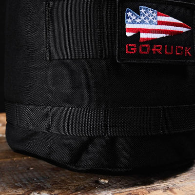 A close-up shot of the Brick Bag (5-50 LBS), a black fabric bag from GORUCK, designed as versatile training gear. It features a patch showcasing a spearhead design with the American flag, with "GORUCK" embroidered in red beneath it. Resting on a wooden surface, this military gear bag embodies durability and style.