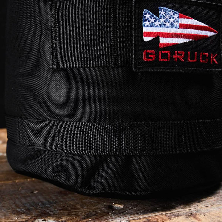 A close-up shot of the Brick Bag (5-50 LBS), a black fabric bag from GORUCK, designed as versatile training gear. It features a patch showcasing a spearhead design with the American flag, with "GORUCK" embroidered in red beneath it. Resting on a wooden surface, this military gear bag embodies durability and style.