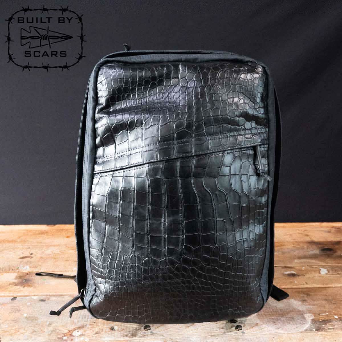 A sophisticated black backpack featuring an alligator leather front panel stands upright on a wooden surface, reminiscent of GORUCK craftsmanship. In the top left corner, the phrase "Built by Scars" appears alongside a fish logo.