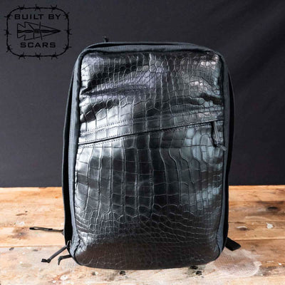 A sophisticated black backpack featuring an alligator leather front panel stands upright on a wooden surface, reminiscent of GORUCK craftsmanship. In the top left corner, the phrase "Built by Scars" appears alongside a fish logo.