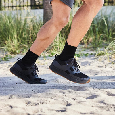 Wearing the Men's Rough Runner in Black + Gum by GORUCK, a person jogs on a sandy beach. The sneakers feature a gradient density EVA midsole and are paired with black socks and gray shorts. Lush greenery and a partial view of a fence in the background capture the essence of a warm, sunny day.
