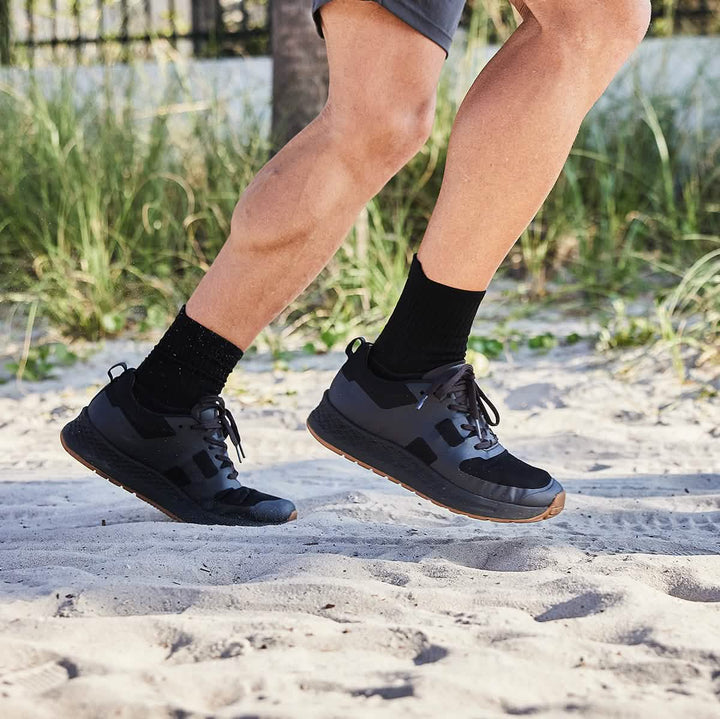 Wearing the Men's Rough Runner in Black + Gum by GORUCK, a person jogs on a sandy beach. The sneakers feature a gradient density EVA midsole and are paired with black socks and gray shorts. Lush greenery and a partial view of a fence in the background capture the essence of a warm, sunny day.