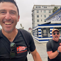 Two men smile and walk outdoors near a beach club with a striped blue and white building, both wearing the comfortable Arnold Pump Club Tee - Tri-Blend by GORUCK.