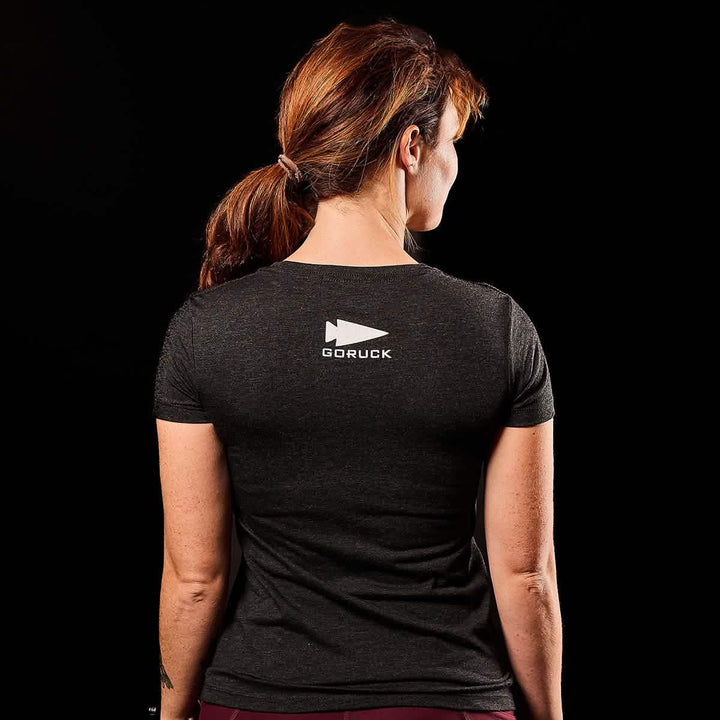 A woman wearing a "Women's CrossFit Tee - Tri-Blend" by GORUCK is facing backwards against a black background.