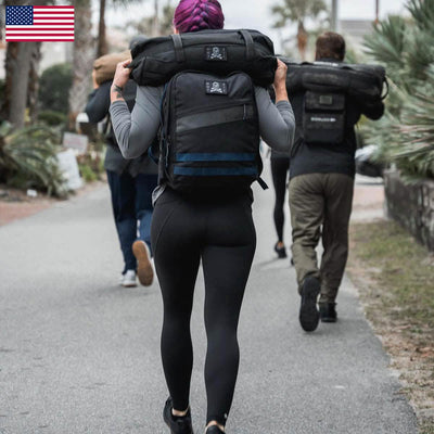 A group of people trudges along a path, each shouldering heavy sandbags. In the foreground, someone with purple hair sports squat-proof GORUCK Women's Training Leggings Pocket made with ToughFlex Fabric. They wear a gray top under the ever-watchful gaze of an American flag in the top left corner.