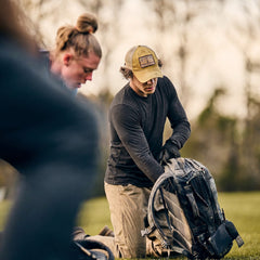 Two people outdoors kneeling on grass, one packing a backpack, both focused while wearing Men’s Slick Long Sleeve - Tri-BlendX shirts made from premium tri-blend fabric with antimicrobial treatment for extra comfort.