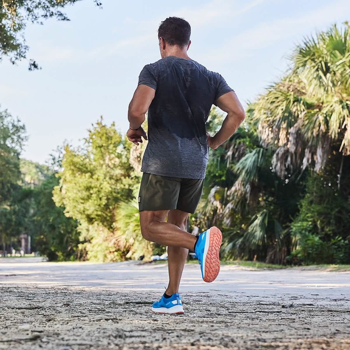A man is jogging along a sunlit path enveloped by lush greenery, exemplifying the essence of fitness and nature. Wearing GORUCK's Men's Rough Runner - Electric Blue shoes with orange soles, paired with a dark gray shirt and olive shorts, he truly embodies the spirit of an outdoor enthusiast.