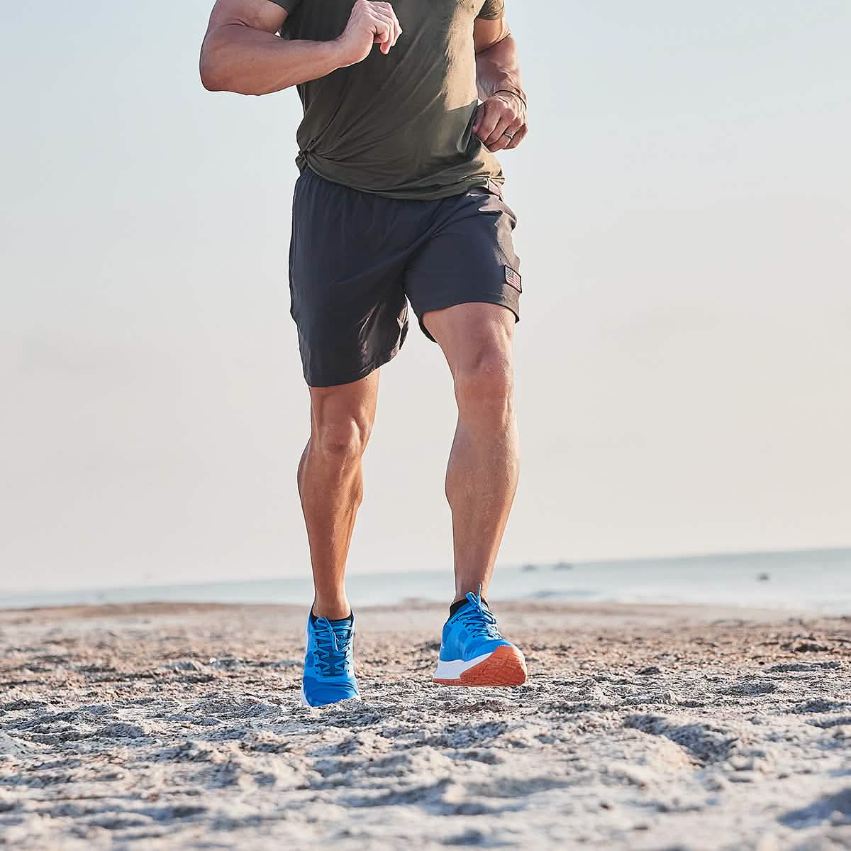 A person jogging on the beach, clad in a green shirt and dark shorts, wearing the Men's Rough Runner - Electric Blue from GORUCK. The Rough Runner enjoys the clear sky as the ocean graces the background with its serene presence.