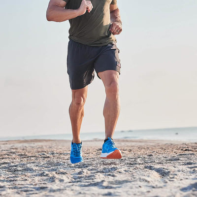 A person jogging on the beach, clad in a green shirt and dark shorts, wearing the Men's Rough Runner - Electric Blue from GORUCK. The Rough Runner enjoys the clear sky as the ocean graces the background with its serene presence.