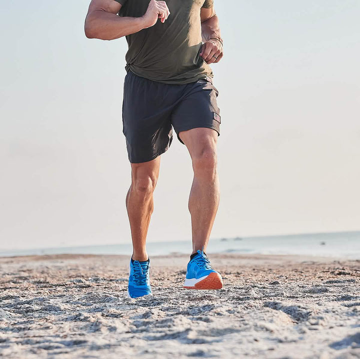 A person jogging on the beach, clad in a green shirt and dark shorts, wearing the Men's Rough Runner - Electric Blue from GORUCK. The Rough Runner enjoys the clear sky as the ocean graces the background with its serene presence.