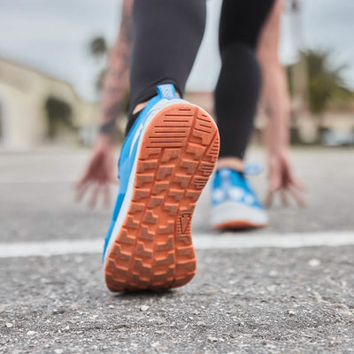 A person in running gear is in a crouched starting position on an outdoor track, highlighting their GORUCK Men's Rough Runner shoes in Electric Blue. These high mileage running shoes feature a textured sole and Gradient Density EVA midsole, engineered for endurance and ideal for the rough runner's journey.