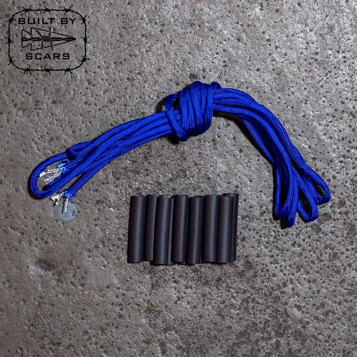 Neatly coiled next to a row of cylindrical black objects on a textured concrete surface is a paracord rope. In the upper left corner, an emblem with "Built By SCARS" is visible, highlighting the ideal setting for DIY ruck upgrades or crafting with the Paracord Kit by GORUCK.