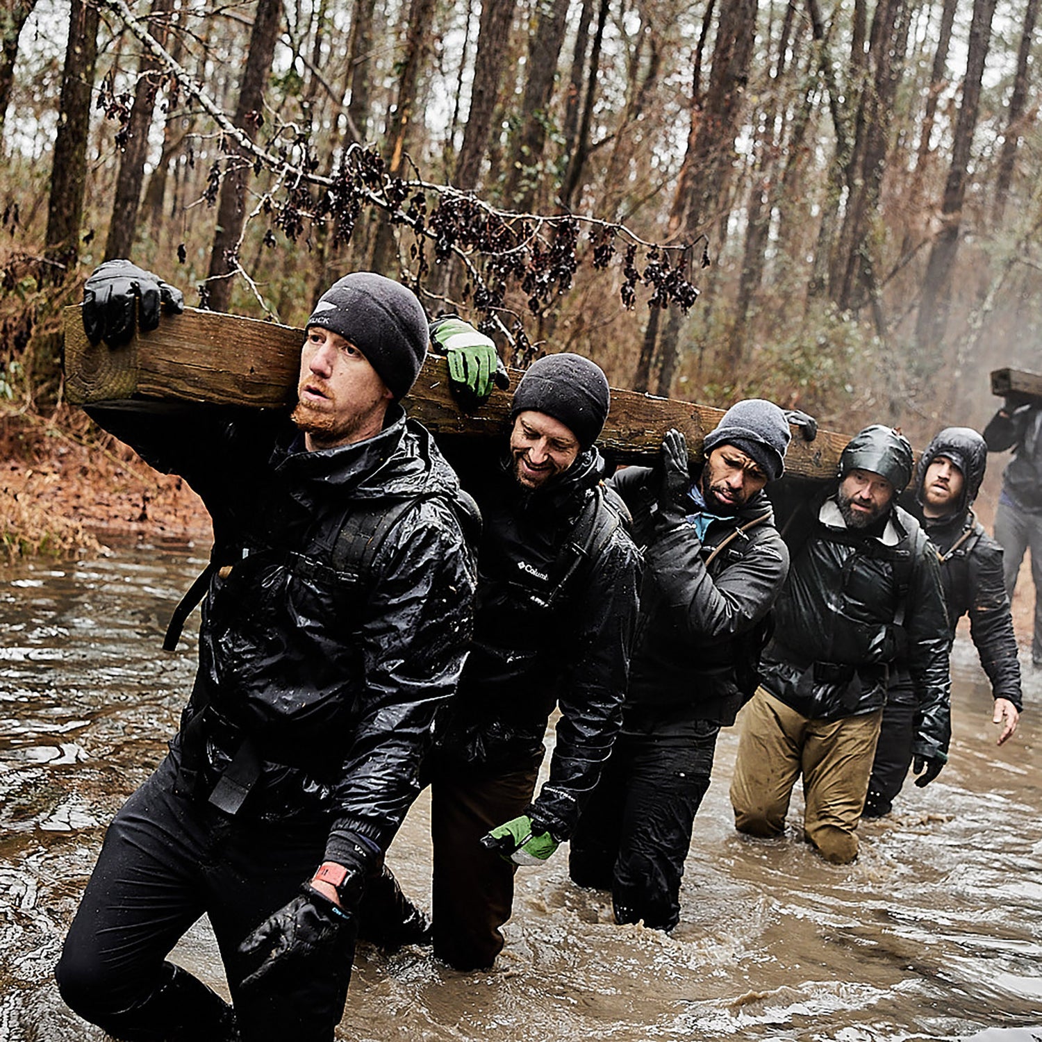 Five people in cold-weather gear carry a log through a muddy, flooded forest during an outdoor challenge.