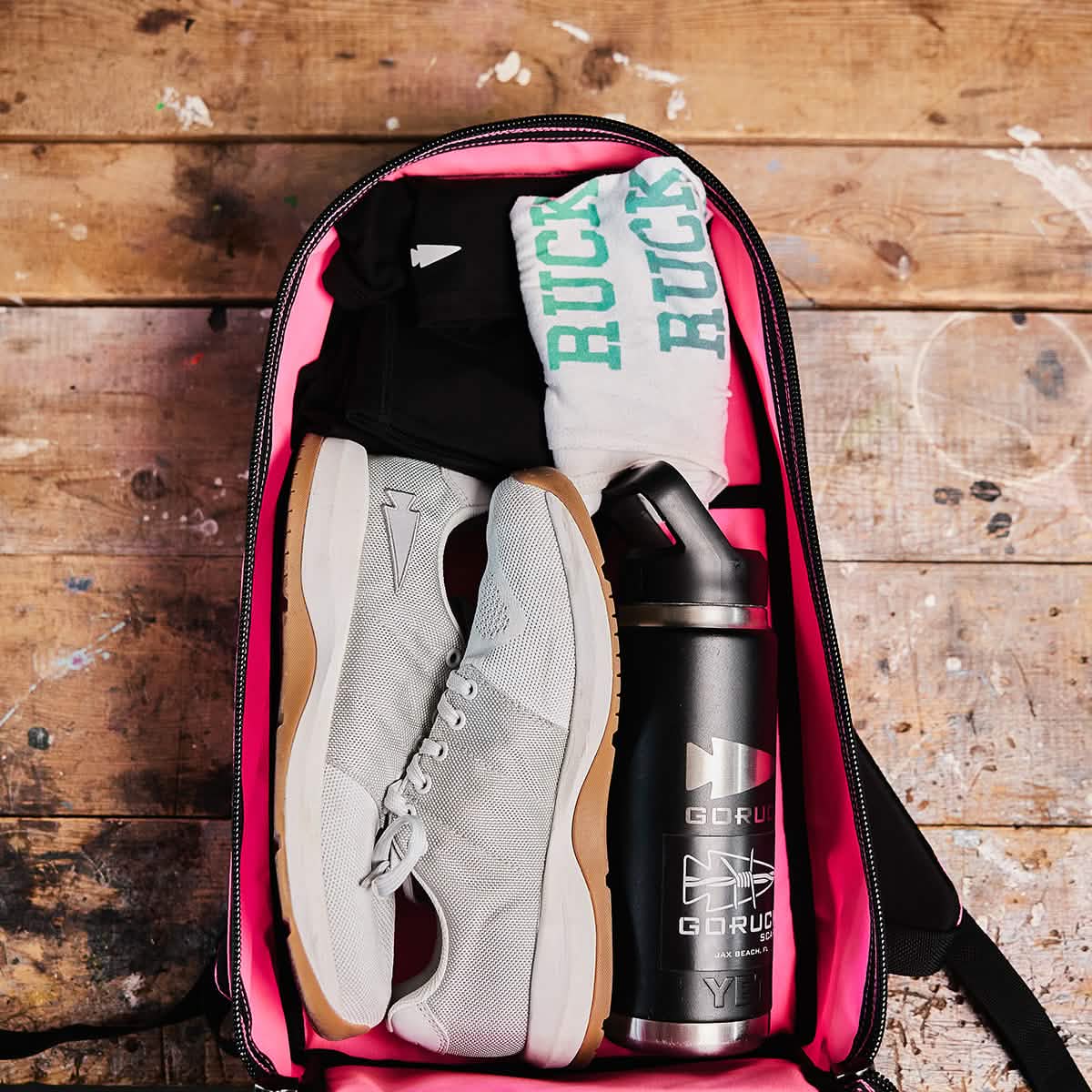 The Bullet Ruck - Classic - Hot Pink gym bag, crafted from rugged Cordura, holds sneakers, socks, a shirt, and a water bottle on a wooden surface—ideal for rucking or use as your daily daypack.
