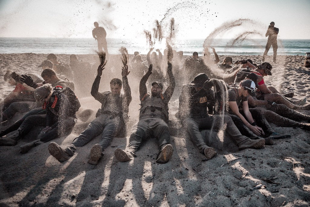 A group of people sit on a sandy beach, tossing sand into the air with both hands.