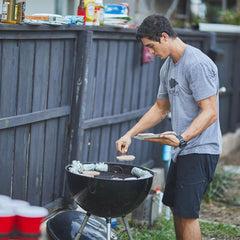 Man in gray GORUCK t-shirt grilling burgers and foil-wrapped food outdoors on charcoal grill