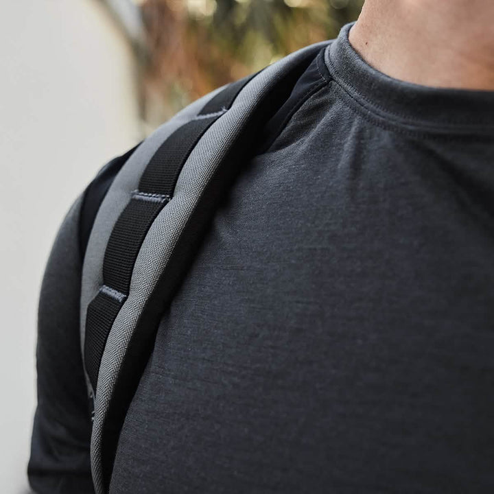 A close-up of a person wearing the Men’s Commando Long Sleeve in Merino Wool by GORUCK, featuring a dark shirt with reinforced paneling and a black and gray backpack strap, standing outside against greenery with a white wall in the background.