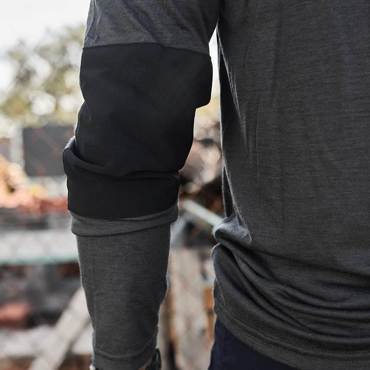 Standing outside, a person is wearing the GORUCK Men’s Commando Long Sleeve made from Merino Wool. The shirt is dark gray with black padded elbow patches reminiscent of a classic British Commando Sweater. The background is blurred, with hints of sunlight and fencing.