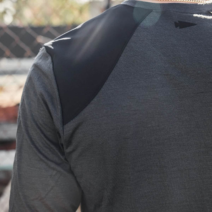 Close-up of a person wearing the Men’s Commando Long Sleeve - Merino Wool by GORUCK, featuring a dark gray athletic design with black shoulder accents, standing outdoors. Sunlight creates soft shadows on the fabric, and a chain-link fence is blurred in the background.