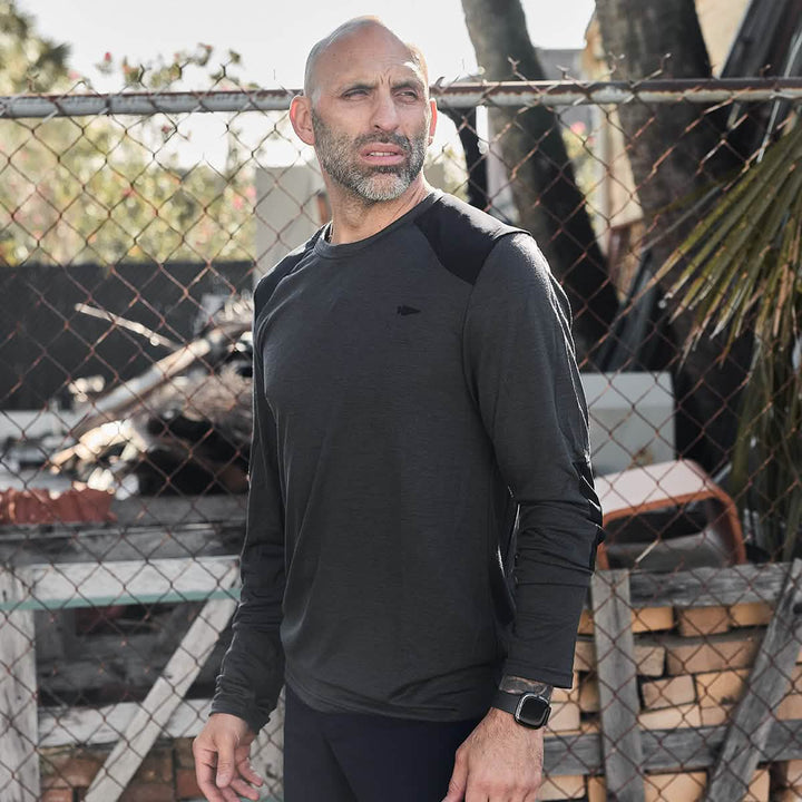 A man with a beard stands outdoors in front of a chain-link fence, wearing a dark Men’s Commando Long Sleeve - Merino Wool by GORUCK and a smartwatch. Sunlight filters through nearby trees, casting shadows on the ground.