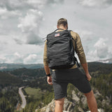 Man wearing a GORUCK backpack with American flag patch, overlooking a scenic mountain landscape.