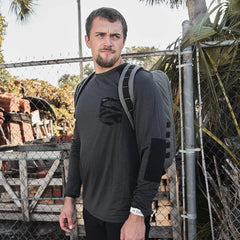 Man wearing charcoal GORUCK backpack and long sleeve shirt standing near chain-link fence with palm trees