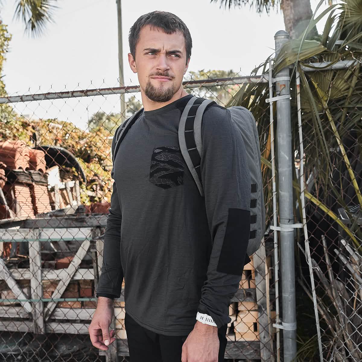 A person is standing outdoors by a chain-link fence, wearing the GORUCK Men’s Commando Long Sleeve in soft merino wool and carrying a backpack. In the background, there are plants and stacked materials as they gaze off to the side.