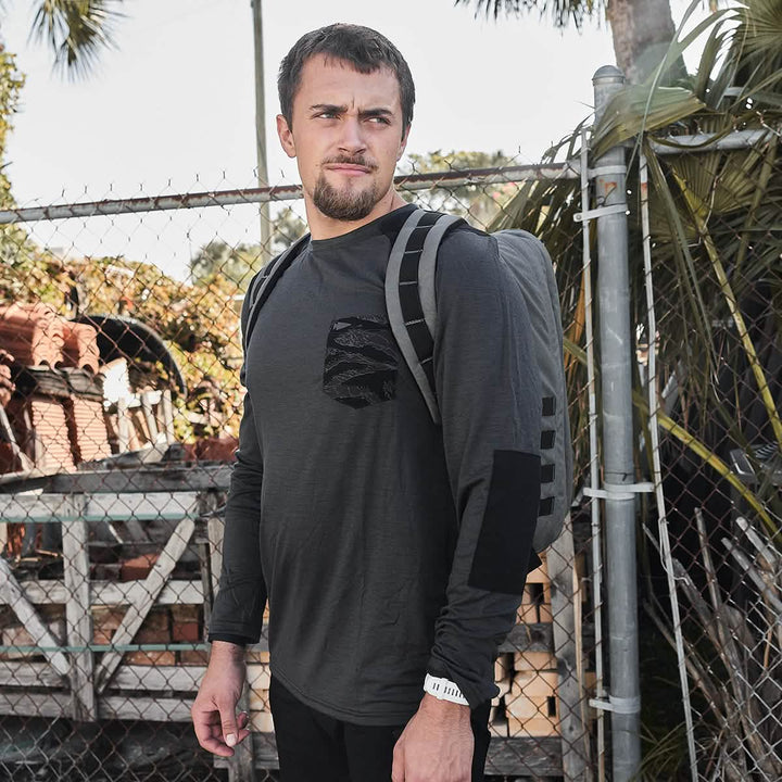 A person is standing outdoors by a chain-link fence, wearing the GORUCK Men’s Commando Long Sleeve in soft merino wool and carrying a backpack. In the background, there are plants and stacked materials as they gaze off to the side.