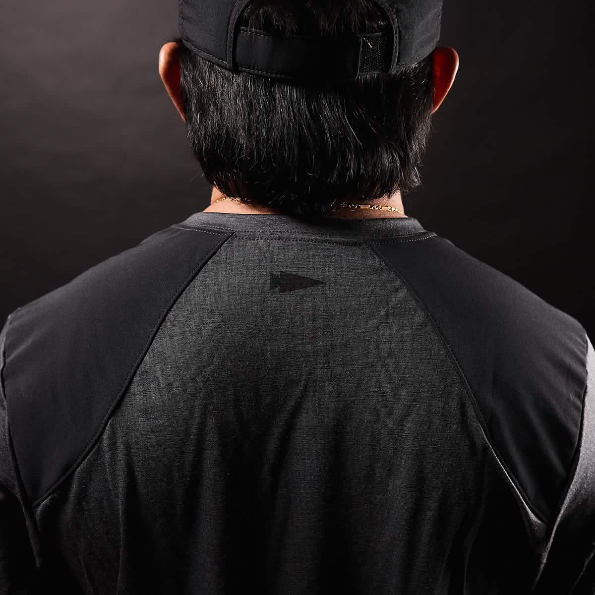 The image depicts the back of a person wearing a black cap and the GORUCK Men's Commando Long Sleeve - Merino Wool, which has a dark, textured appearance similar to a British Commando Sweater. The emphasis is on the upper back, showcasing reinforced paneling with a subtle geometric logo at its center. The background features a dark gradient.