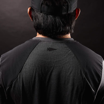 The image depicts the back of a person wearing a black cap and the GORUCK Men's Commando Long Sleeve - Merino Wool, which has a dark, textured appearance similar to a British Commando Sweater. The emphasis is on the upper back, showcasing reinforced paneling with a subtle geometric logo at its center. The background features a dark gradient.