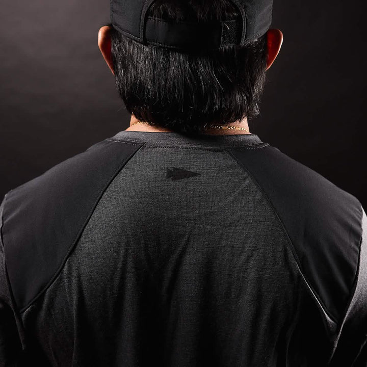 The image depicts the back of a person wearing a black cap and the GORUCK Men's Commando Long Sleeve - Merino Wool, which has a dark, textured appearance similar to a British Commando Sweater. The emphasis is on the upper back, showcasing reinforced paneling with a subtle geometric logo at its center. The background features a dark gradient.