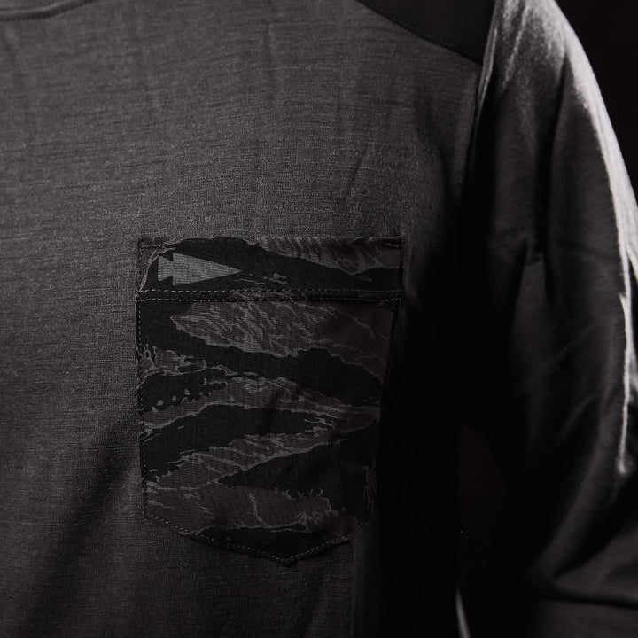 Close-up of the Men’s Commando Long Sleeve - Merino Wool by GORUCK, featuring a dark gray hue with a single front pocket. The pocket showcases a black and gray camo pattern, subtly contrasting with the plain fabric and evoking the rugged style reminiscent of a British Commando Sweater. Displayed against a dark background, the shirt's refined design stands out beautifully.