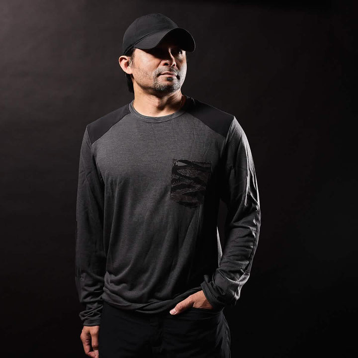 A man wearing the Men’s Commando Long Sleeve - Merino Wool by GORUCK stands against a black background. With his hand in his pants pocket, he gazes to the side as dramatic shadows play across the scene.