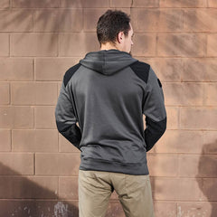 A man stands with his back to the camera, wearing the Men’s Full Zip - Polartec Grid Fleece from GORUCK in a gray color with black accents and khaki pants. He is positioned in front of a textured, light brown brick wall that features faint tree branch shadows.
