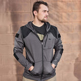 A person stands against a light brown brick wall, wearing the GORUCK Men’s Full Zip - Polartec Grid Fleece jacket in gray and black, with hands in pockets. They have short hair and a beard, gazing to the side as soft shadows from the sun fall on the wall.