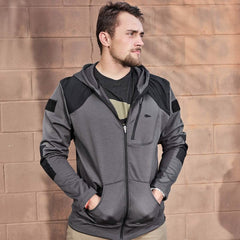 A person stands against a light brown brick wall, wearing the GORUCK Men’s Full Zip - Polartec Grid Fleece jacket in gray and black, with hands in pockets. They have short hair and a beard, gazing to the side as soft shadows from the sun fall on the wall.