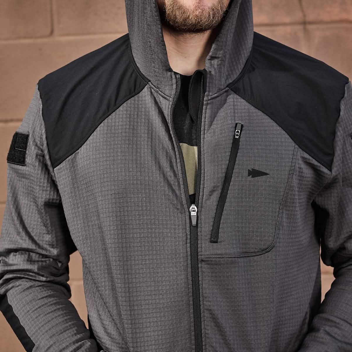 A person in a gray and black GORUCK Men’s Full Zip - Polartec Grid Fleece jacket, complete with a hood, stands against a brown brick wall. The jacket highlights its Polartec Power Grid fabric, boasting a zippered chest pocket and adorned with a small emblem on the front.