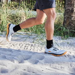 A person running on a sandy beach in Mackall trail shoes by GORUCK, featuring the Glacier + Ignition Orange colorway and a triple compound outsole, along with black socks and dark shorts. Tall grass and a fence are visible in the background.