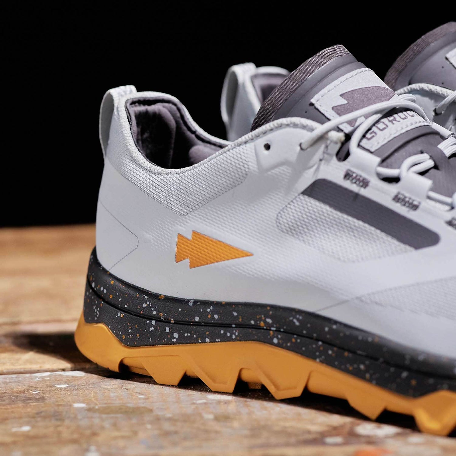 A close-up of the Mackall - Glacier + Ignition Orange athletic shoes by GORUCK, featuring white and gray tones with striking orange accents, specifically designed for Special Forces training. These rucking shoes boast a speckled black midsole and a rugged triple compound orange outsole, all elegantly displayed on a wooden surface.