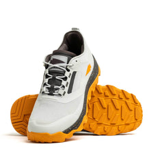 A pair of the Mackall by GORUCK in Glacier + Ignition Orange features stylish trail running shoes with white mesh uppers and vibrant orange soles. One shoe stands on its side, highlighting the rugged triple compound outsole pattern for enhanced grip.