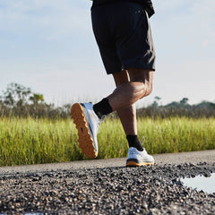 A person wearing GORUCK's Mackall - Glacier + Ignition Orange, featuring a triple compound outsole, jogs on a rural road. The focus is on their legs and footwear. Lush green fields and trees extend into the distance, while a puddle reflects the sky on the asphalt.