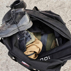 An open black GORUCK Kit Bag (Includes Shoulder Strap) rests on the ground, revealing clothing, a pair of black boots, and various items tucked inside. The slightly worn look adds to its charm, hinting at adventures taken.