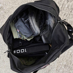 Open a black GORUCK Kit Bag (includes shoulder strap) labeled as a Special Forces Flight Bag, revealing packed items including black boots, folded clothes, and a rolled-up belt marked #001 on a concrete surface.