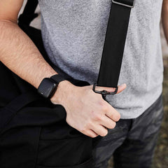 A person in a gray t-shirt and camo pants clutches the shoulder strap of a sleek black GORUCK Kit Bag. A smartwatch adorns their left wrist, capturing attention along with the hand firmly gripping the strap.