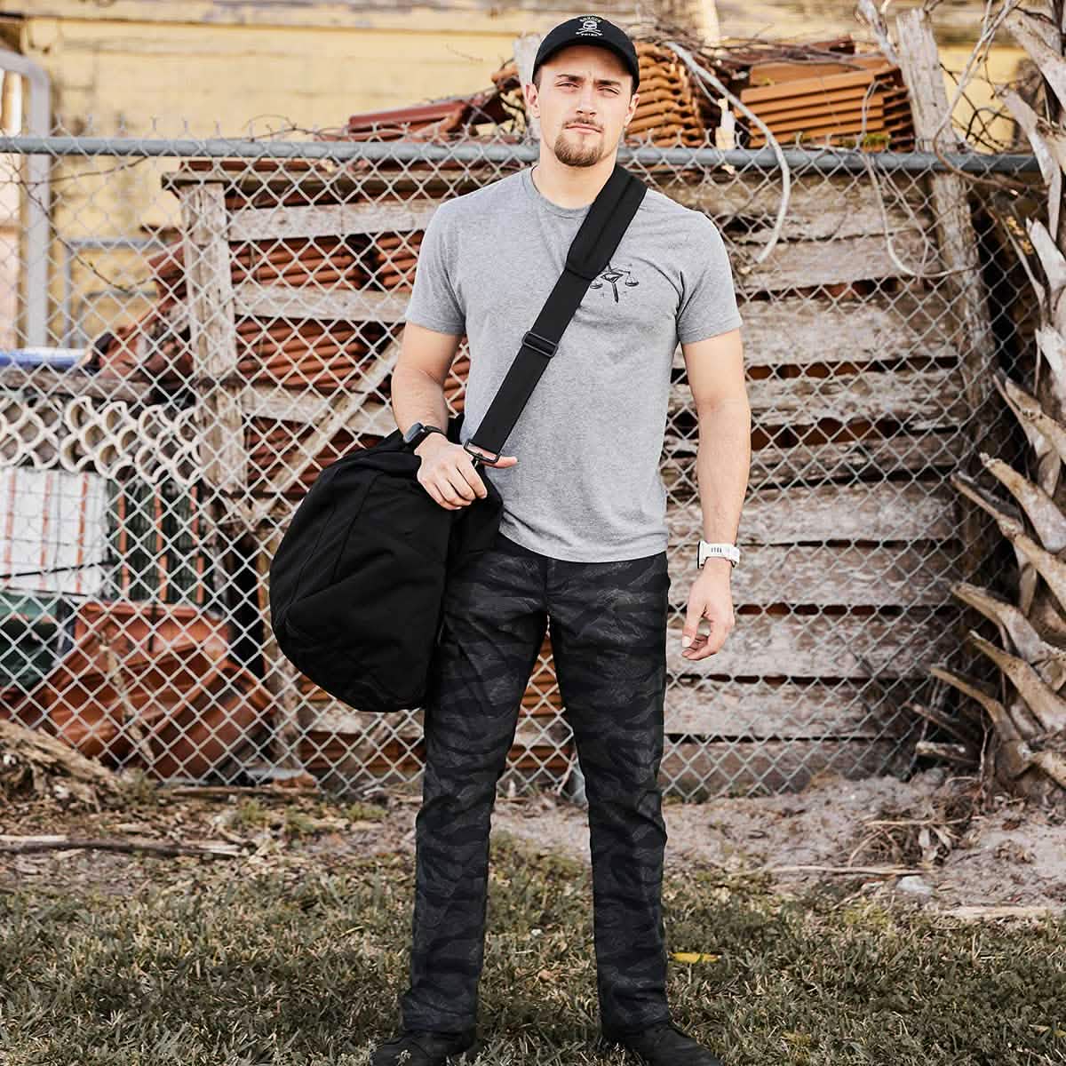 Standing on the grass, a person is dressed in a gray t-shirt, camouflage pants, and a black cap. They are carrying the sleek Kit Bag with a shoulder strap from GORUCK. Behind them is a chain-link fence and stacked wooden pallets.
