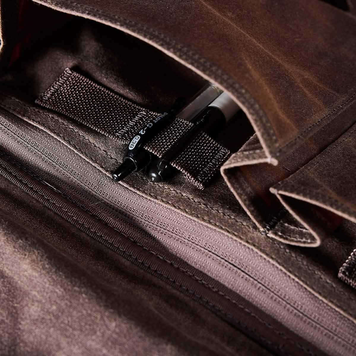 Close-up of an open GORUCK Heritage Claymore Sling Bag crafted from waxed canvas, displaying two black pens inside. The bag highlights a textured fabric lining and includes a zippered pocket. The flap is lifted to reveal the contents, emphasizing its heritage durability.
