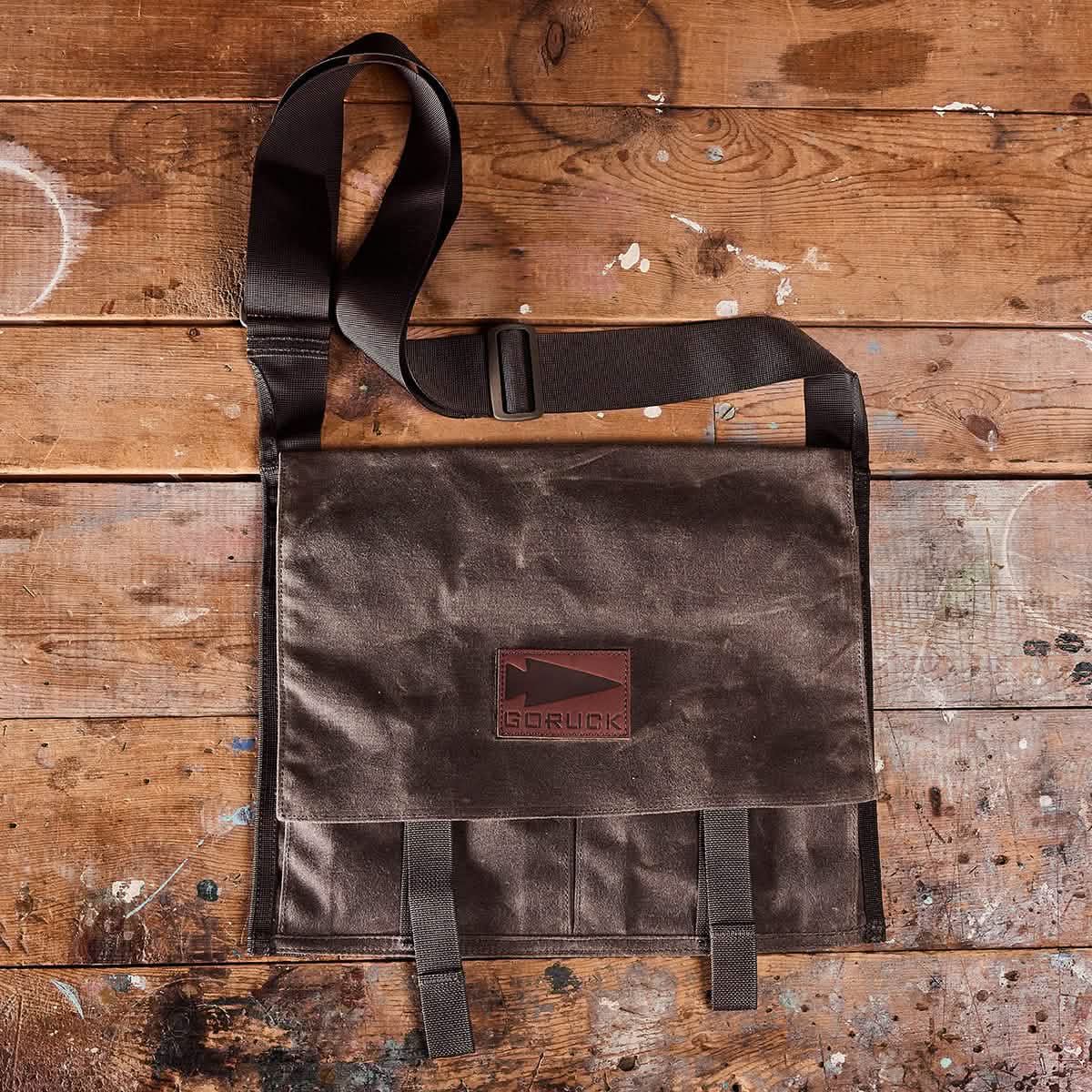 The Heritage Claymore Sling Bag from GORUCK, featuring a brown waxed canvas construction for lasting heritage durability and an adjustable black strap, rests flat on a rustic wooden surface. The bag is adorned with a rectangular logo patch centered on its face. The background wood surface bears paint splatters and shows signs of wear for a distressed look.