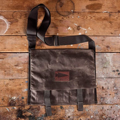 The Heritage Claymore Sling Bag from GORUCK, featuring a brown waxed canvas construction for lasting heritage durability and an adjustable black strap, rests flat on a rustic wooden surface. The bag is adorned with a rectangular logo patch centered on its face. The background wood surface bears paint splatters and shows signs of wear for a distressed look.