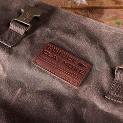 Close-up of a brown Heritage Claymore Sling Bag by GORUCK, featuring a leather patch with "LIFETIME GUARANTEE" inscribed. Crafted from waxed canvas for heritage durability, it has dark straps with metal buckles, displayed against a wooden surface with green paint splatters.