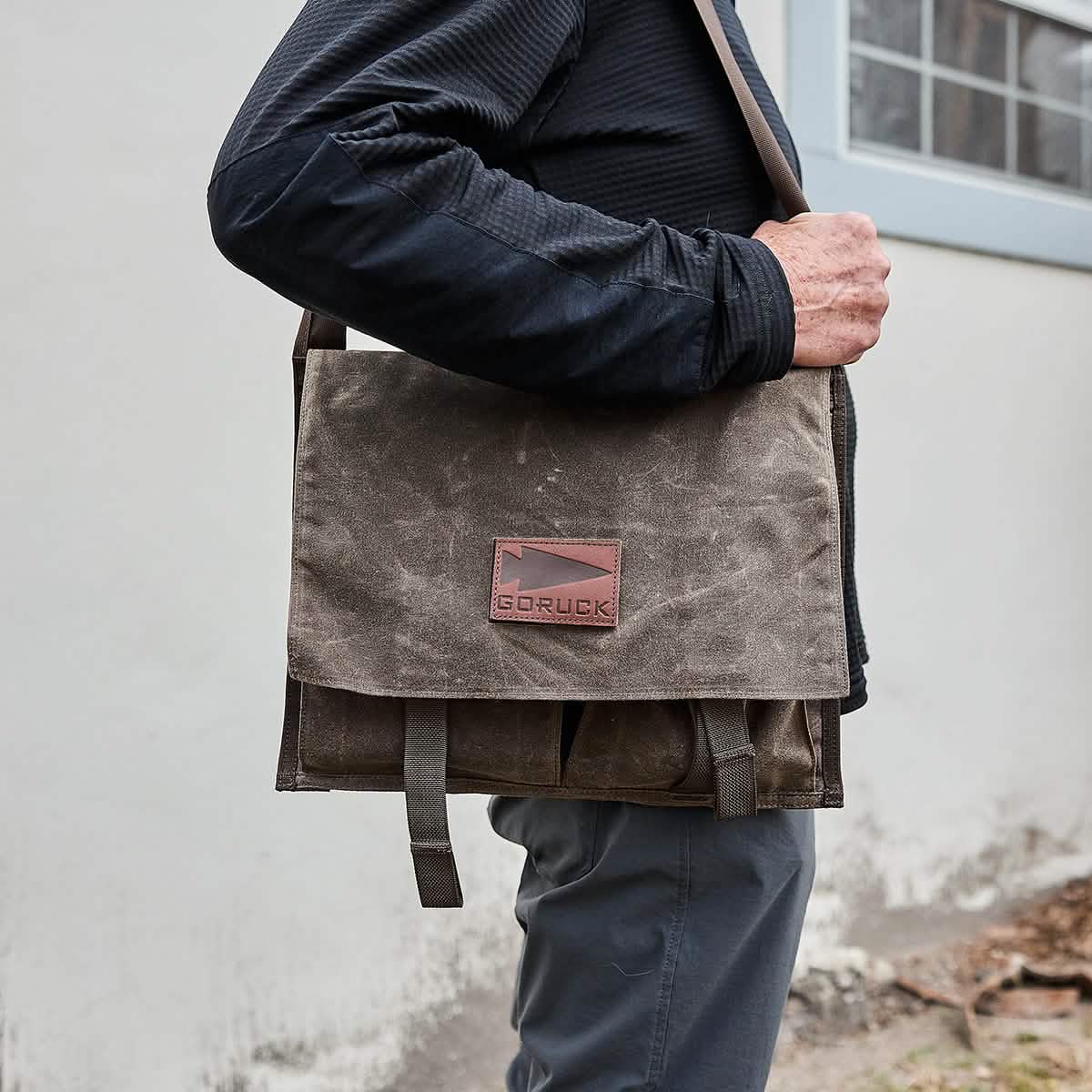 A person in a black jacket and gray pants is holding a brown GORUCK Heritage Claymore Sling Bag, demonstrating its timeless durability. The bag has two straps and is worn over the shoulder. The background features a white wall and a window.